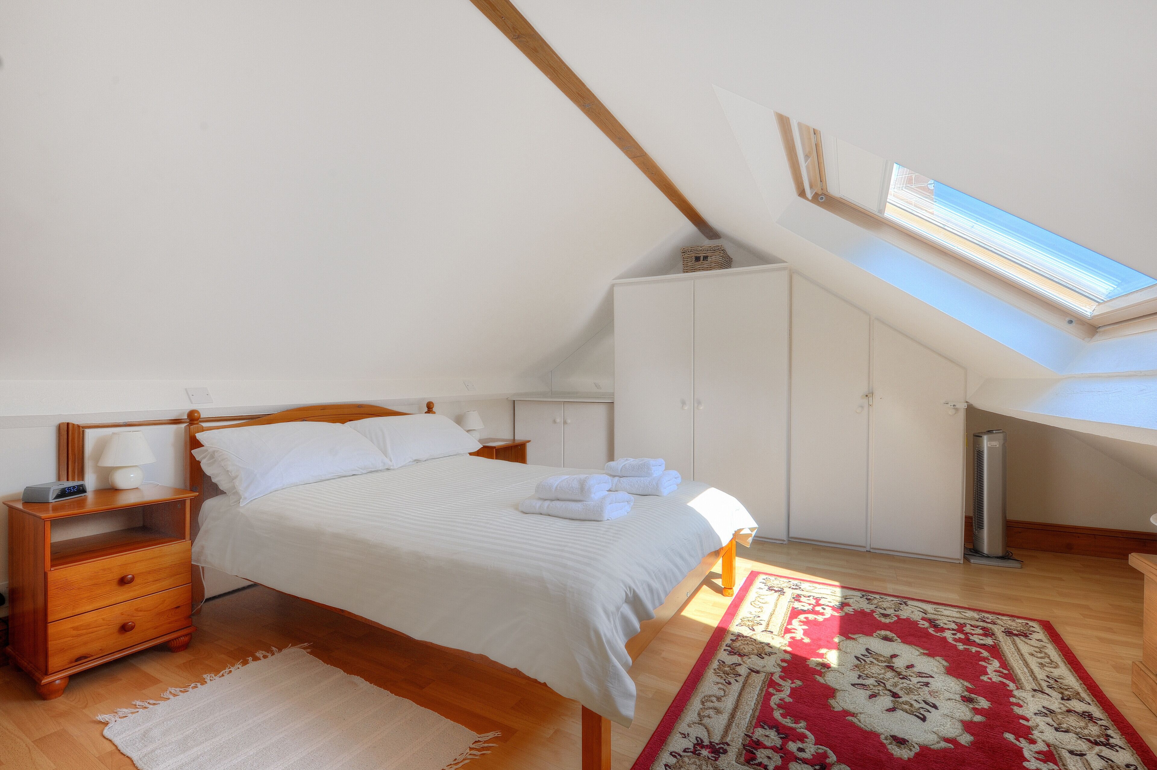 A bedroom with a sloped ceiling, a skylight, a bed with white linens, a wooden nightstand, a red and white patterned rug, and a white wardrobe.