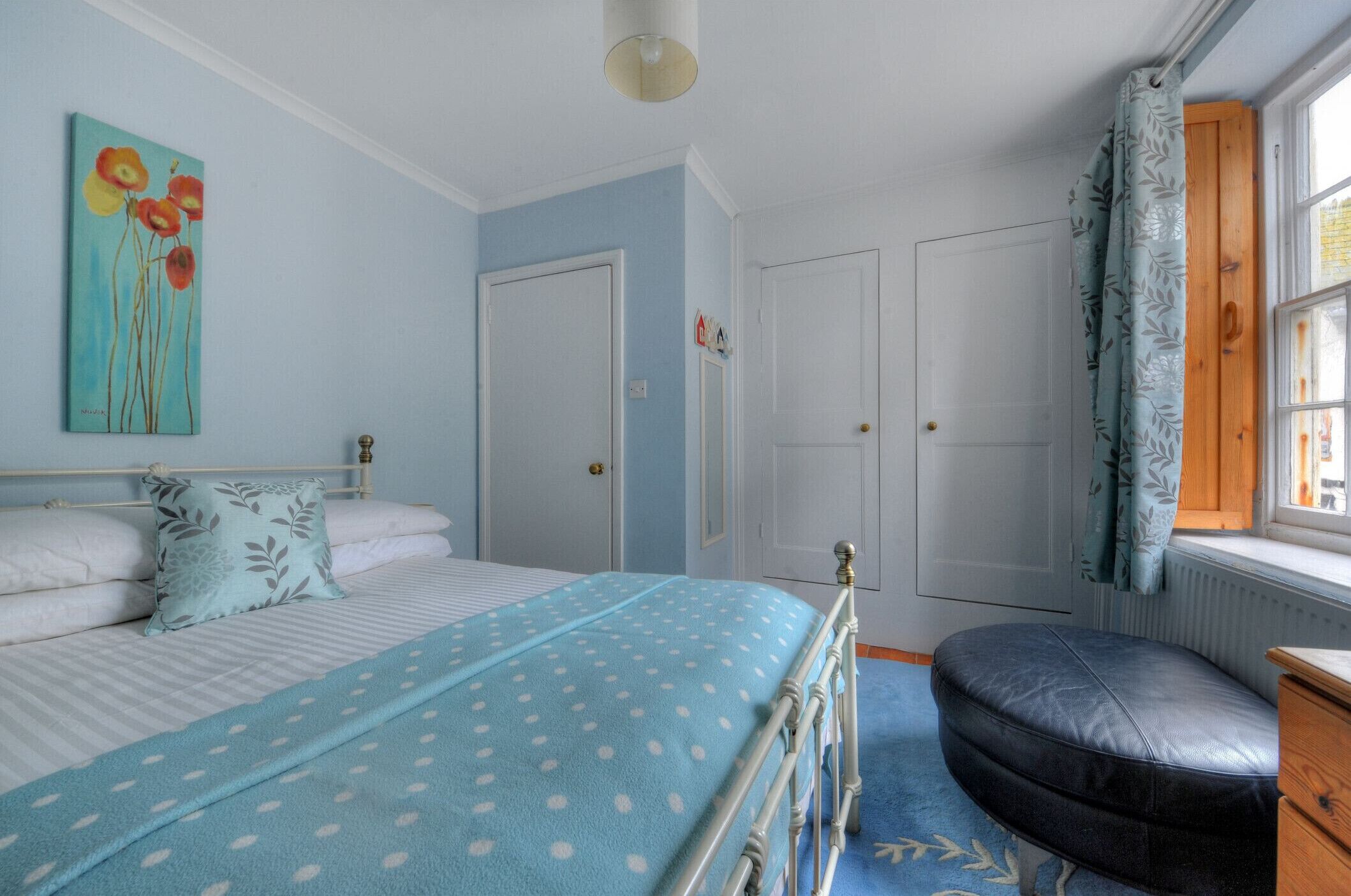 A bedroom with a bed, a blue polka dot blanket, a pillow, a wooden bench, and a window with curtains.