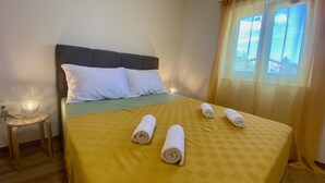 Ein ordentlich bezogenes Bett mit gelber Tagesdecke und weißen Kissen, ein kleiner Nachttisch mit Lampe und ein Fenster mit Vorhängen.