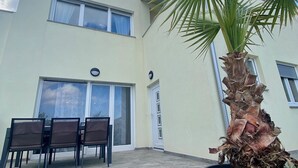 Ein modernes Gebäude mit flachem Dach, mehreren Fenstern und einer Tür. Vor dem Gebäude befindet sich eine Terrasse mit Tisch, Stühlen und einem Blumentopf.