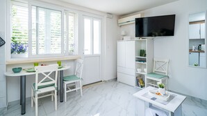 A bright, modern kitchen with white cabinetry, a dining table with chairs, a TV mounted above the cabinets, and a window with a view of greenery. - Apartment Laneta - Studio Apartment with Terrace (Dubrovnik)