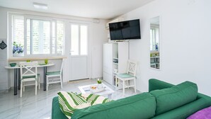 A modern living room with a green sofa, a dining area with a table and chairs, a TV mounted on the wall, and a large window with blinds. - Apartment Laneta - Studio Apartment with Terrace (Dubrovnik)
