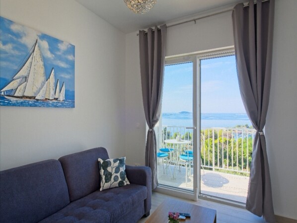 A living room with a sofa, a painting of sailboats, and a view of the sea through a sliding glass door.