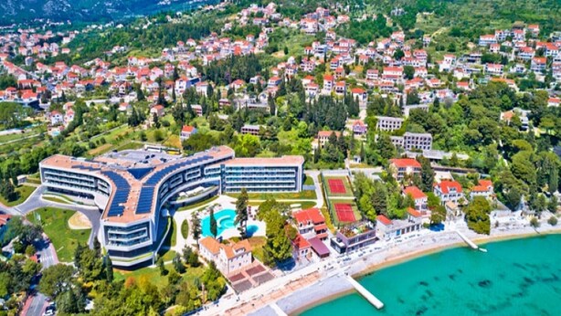 Vista aérea de un pueblo costero con un hotel moderno de diseño curvo, una playa y un puerto deportivo.
