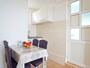 Une cuisine compacte avec des meubles blancs, un pare-feu en carrelage et une table à manger agrémentée d’un panier de fruits.