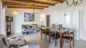 Een moderne woonkamer met een eethoek, houten balken in het plafond, een kroonluchter en een kleurrijk abstract schilderij.
