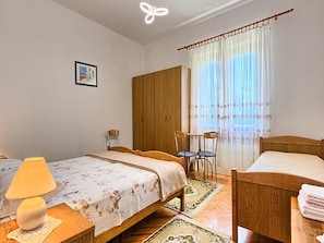 A bedroom with two beds, a wooden wardrobe, a table with a lamp, and a chair.
