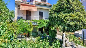 Un edificio de dos pisos con un balcón, rodeado de abundante vegetación y un árbol.
