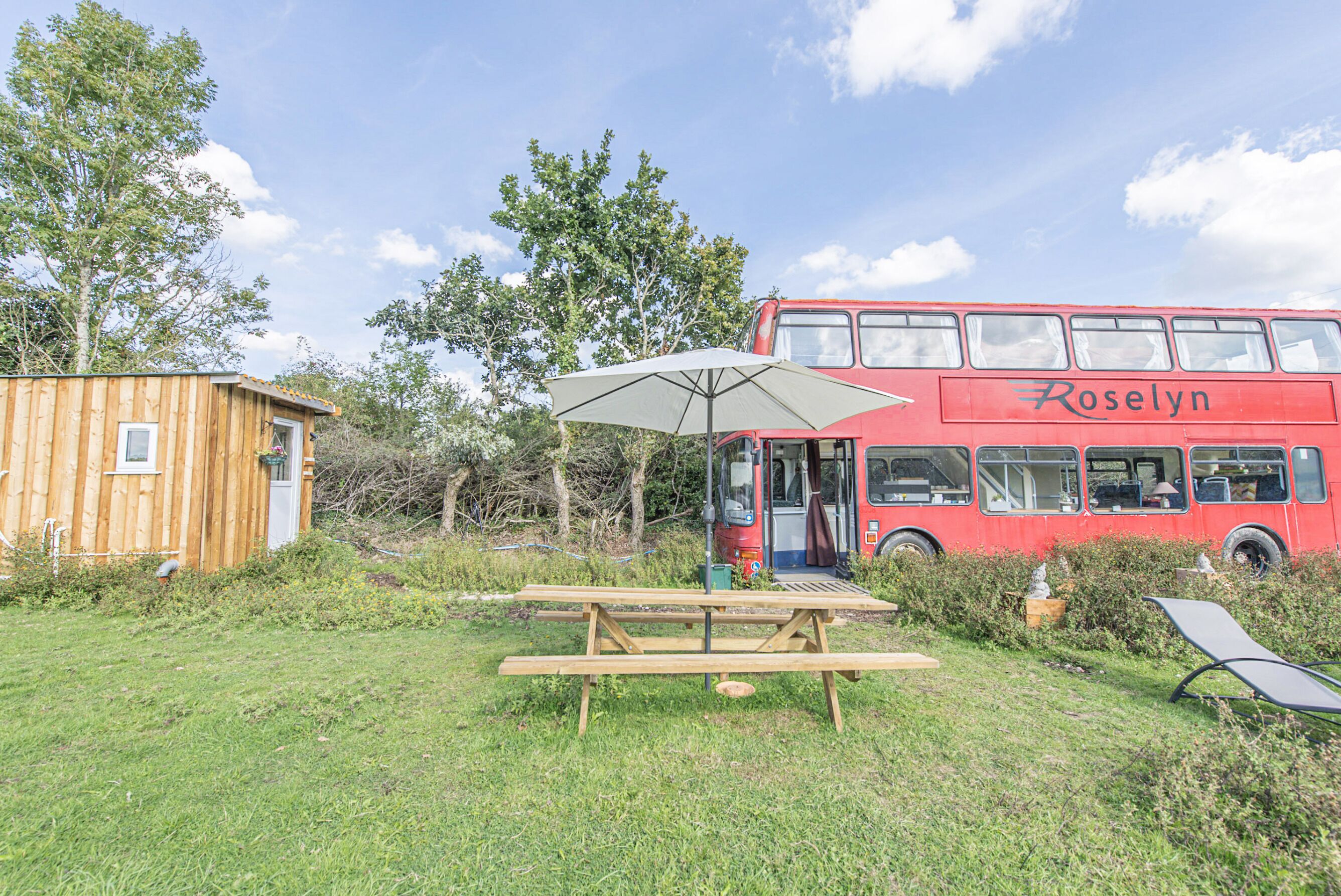 Une table de pique-nique avec un parasol, un autobus à deux étages rouge portant l’inscription « Roselyn », et une structure en bois dans une zone herbeuse.