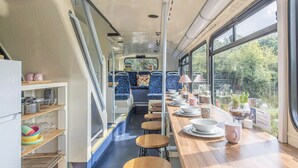 A train dining car with a long wooden table, blue upholstered seats, and a view of greenery outside. - Double Decker Bus on an Alpaca Farm Sleeps 8 (Newton Abbot)