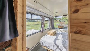A compact, wooden interior with a single bed, a window with curtains, and a view of a green landscape. - Double Decker Bus on an Alpaca Farm Sleeps 8 (Newton Abbot)