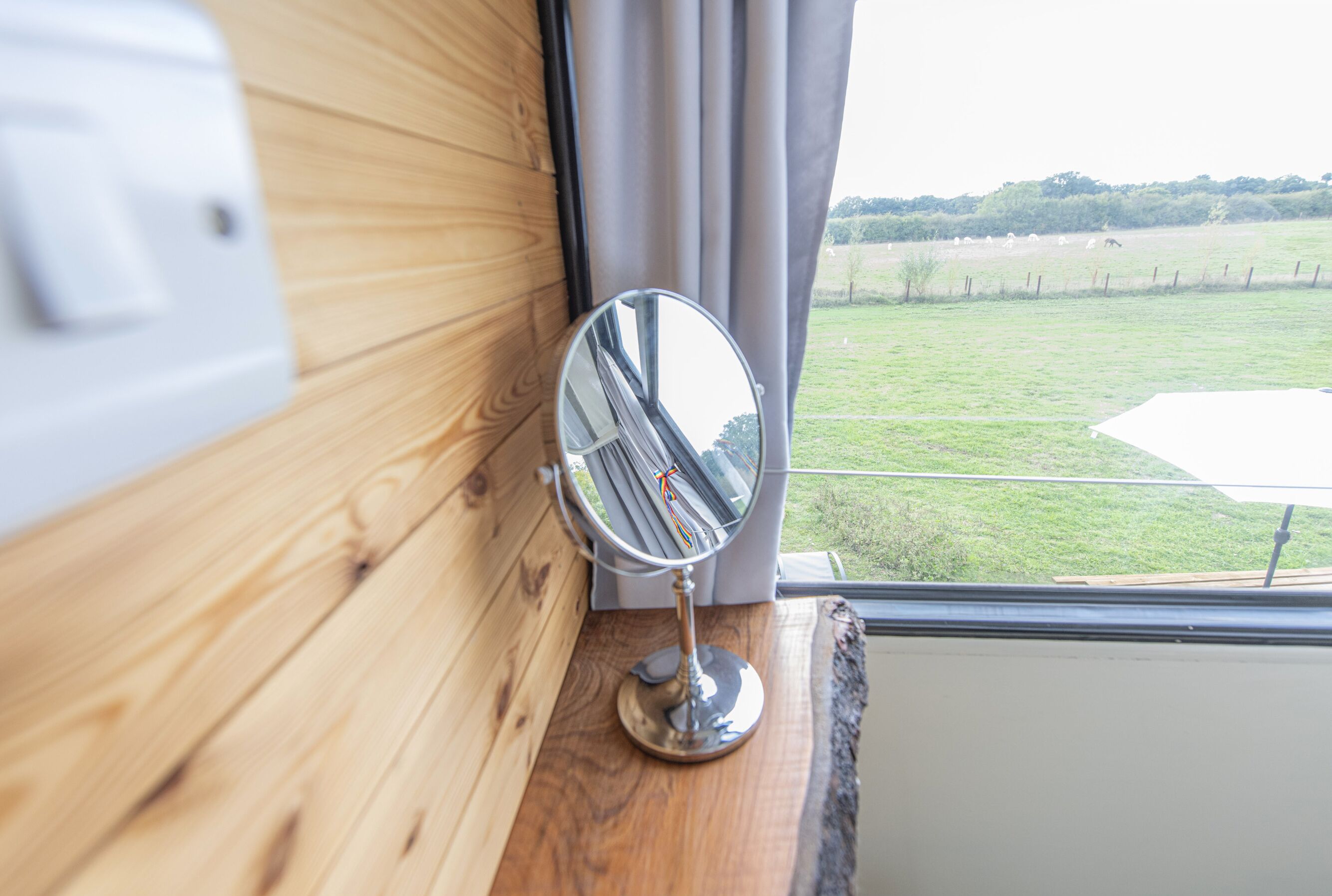 A wooden wall with a mirror reflecting a window view of a grassy field and a fence.