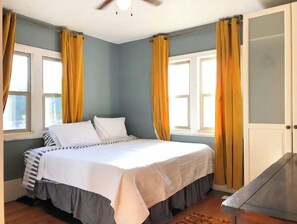 Une chambre à coucher avec un lit, deux fenêtres avec des rideaux, un ventilateur de plafond et une porte.