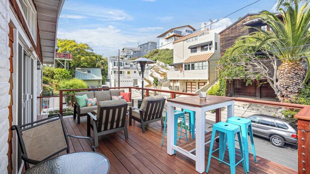 Une terrasse en bois, aménagée avec du mobilier d’extérieur, notamment une table et des chaises, donnant sur une rue bordée de maisons et de verdure.