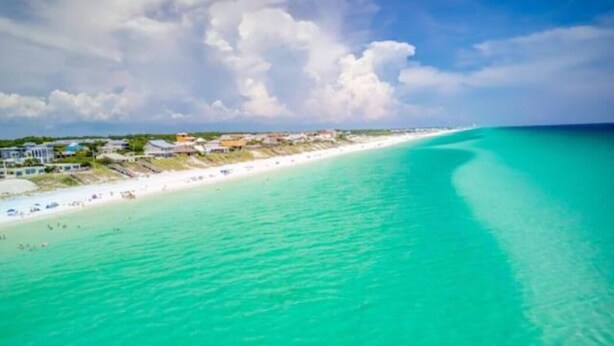 Ein Küstenstrand mit klarem türkisblauem Wasser, einem Sandstrand und Gebäuden im Hintergrund.