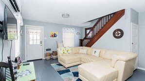 Una sala de estar con un sofá seccional color beige, una escalera de madera, un reloj en la pared y una ventana con persianas.