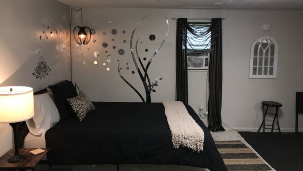 A bedroom with a bed, a nightstand, a lamp, a wall with decorative elements, a window, and a radiator. - Glamping with a twist (Jacksonville)