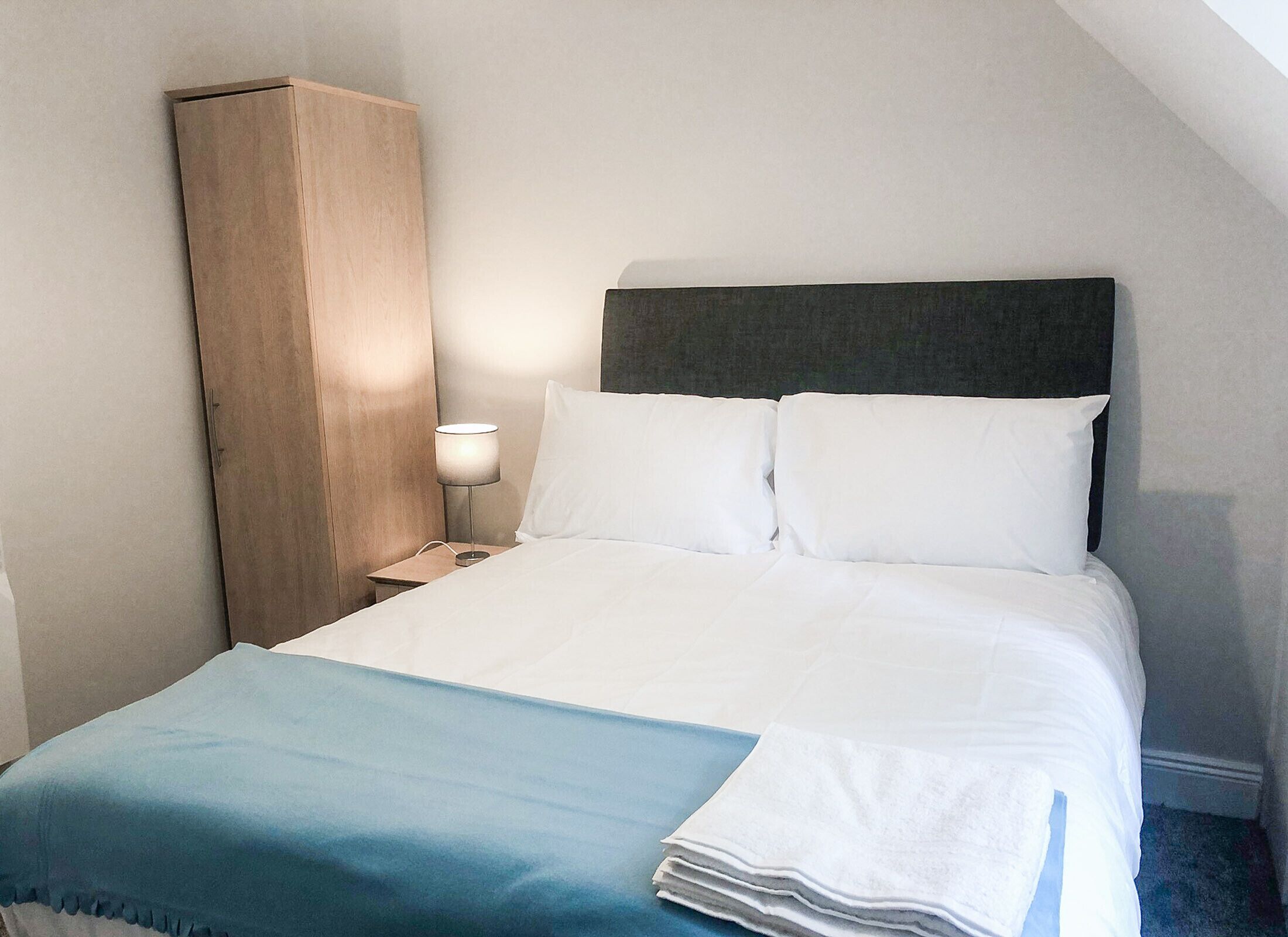 A neatly made bed with white linens and a blue throw blanket, a bedside lamp, and a wooden wardrobe.