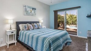 A bedroom with a bed, a nightstand, a ceiling fan, a window with a view of palm trees, and a painting on the wall.