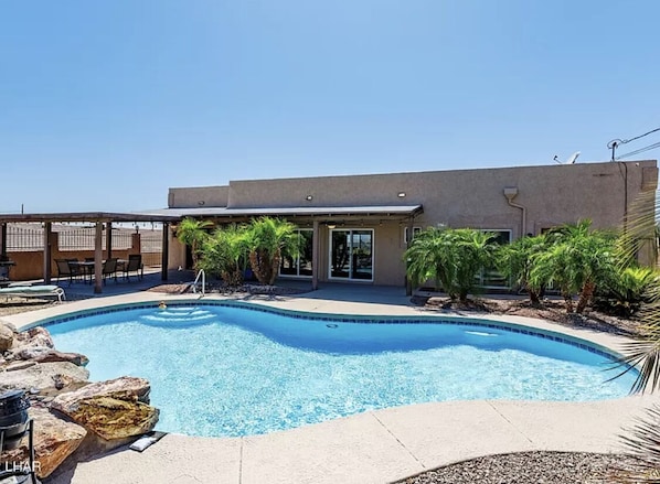 A swimming pool with a modern building in the background, outdoor seating, and palm trees.