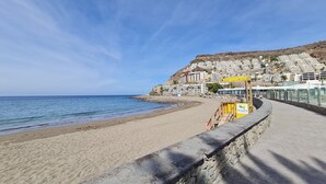 A beach with a lifeguard tower, a rocky cliff, and buildings in the background. - Sunny apartment with sea view (Mogán)