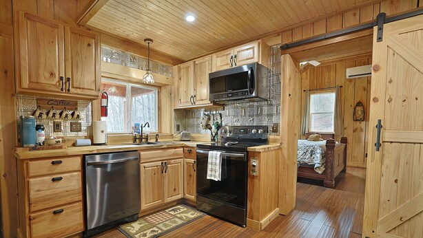 A wooden cabin kitchen with a stainless steel refrigerator, oven, and microwave. There is a wooden dining table and a window with curtains. - Couples Retreat Cabin w HotTub & EV Charger Bryson (Bryson City)