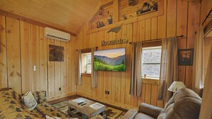 A cozy cabin interior with wooden walls, a mounted TV, a sofa, a chair, and a window with curtains. - Couples Retreat Cabin w HotTub & EV Charger Bryson (Bryson City)