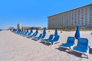Une plage bordée de rangées de chaises longues et de parasols bleus, avec un grand bâtiment d’hôtel en arrière-plan.