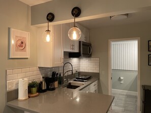 Une cuisine moderne avec des armoires blanches, un évier en acier inoxydable et un four à micro-ondes. Des luminaires suspendus et un tableau encadré ornent le mur.