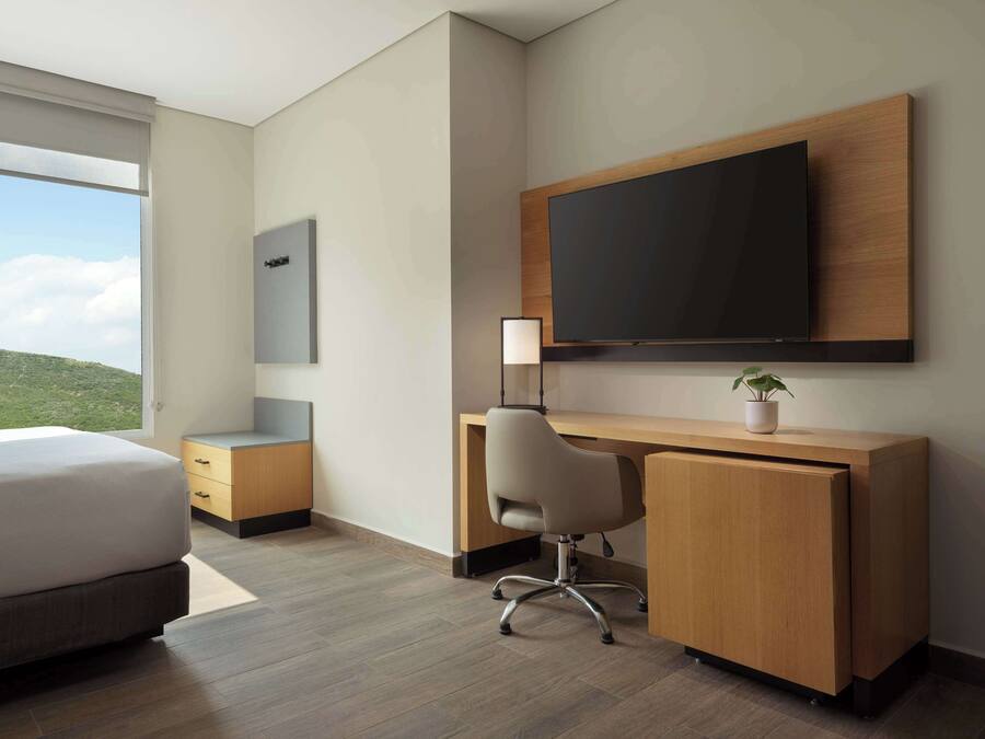 A modern hotel room with a large bed, a wooden desk, a chair, a flat-screen TV mounted on the wall, and a window offering a view of the outdoors.