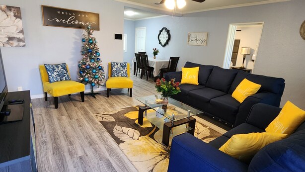 A living room with a Christmas tree, yellow and blue chairs, a black sofa with yellow pillows, a glass coffee table, and a dining area in the background. - Luxurious beds, remodeled, very clean, quiet neighborhood, big backyard & tree!! (Denison)