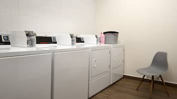 Une buanderie équipée de plusieurs machines à laver et sèche-linge blancs, d’une chaise grise et d’un panier posé sur le comptoir.