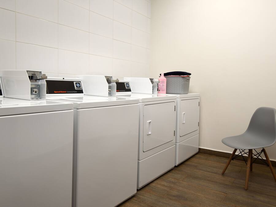Une buanderie équipée de plusieurs machines à laver et sèche-linge blancs, d’une chaise grise et d’un panier posé sur le comptoir.