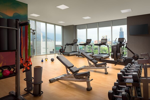 Un gimnasio con equipamiento de ejercicio, ventanas amplias y vistas a la ciudad.