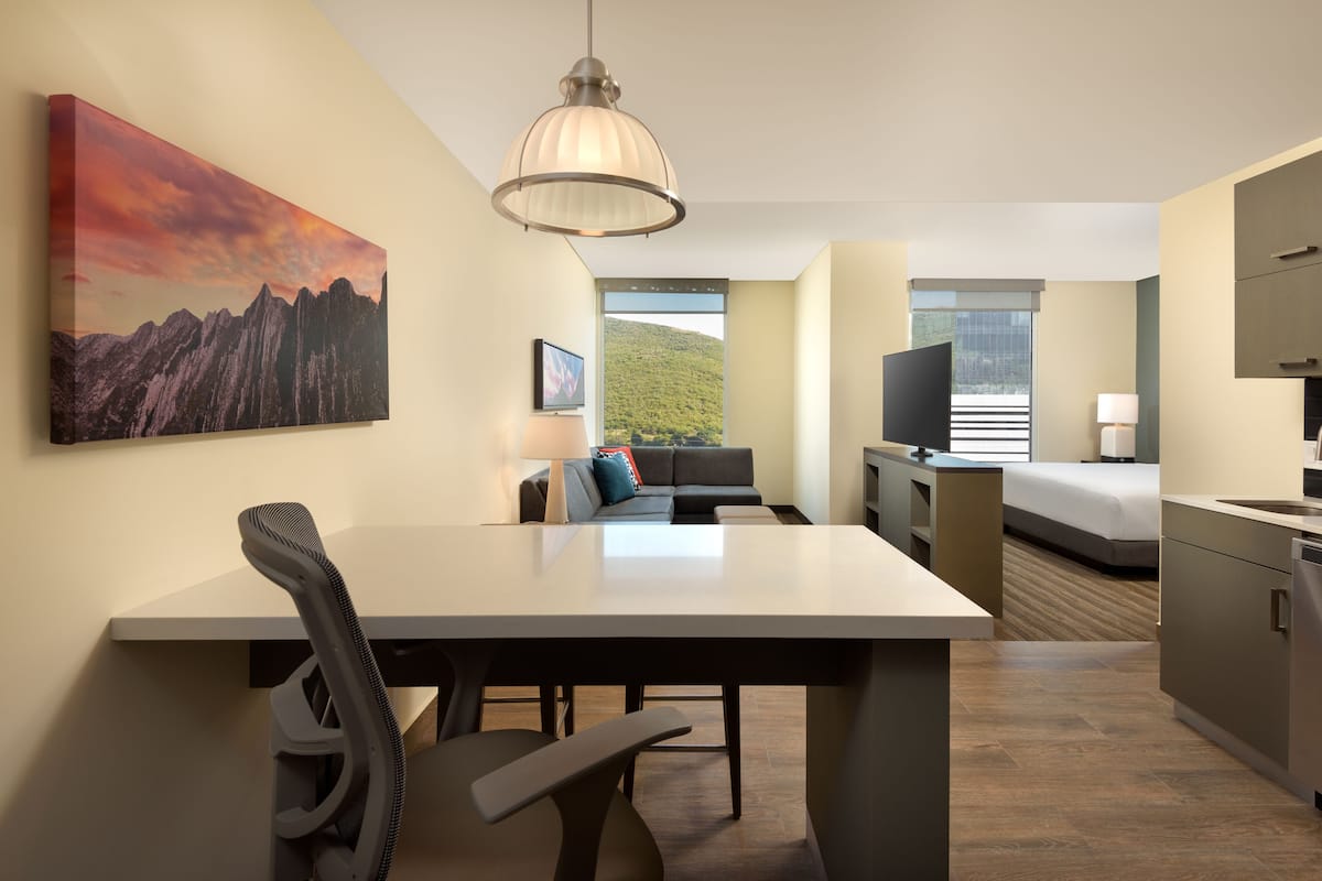 A modern hotel room with a dining area, a sofa, a TV, and a large window with a mountain view.