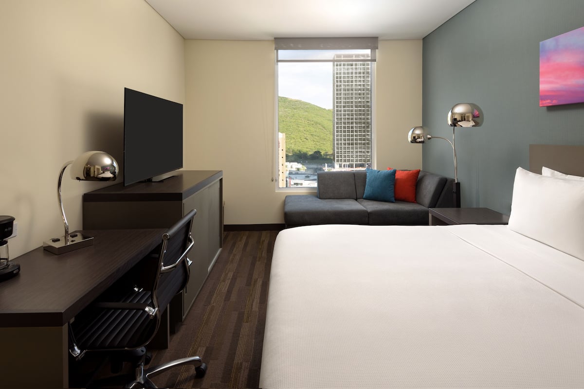 A hotel room with a bed, a desk with a TV, a chair, a sofa with pillows, a lamp, and a window with a view of a building and hill.