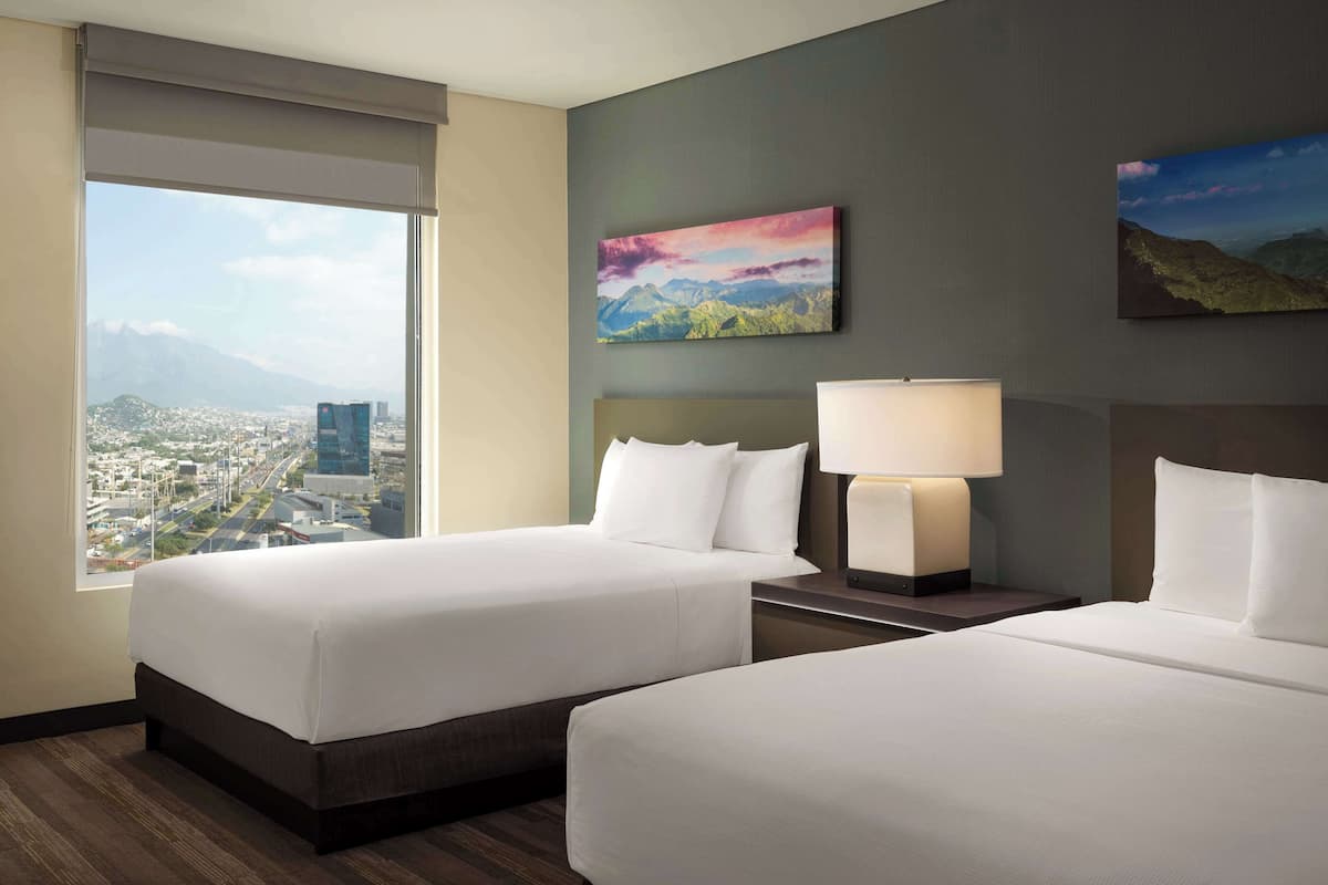 A hotel room with two beds, a city view, and a mountain landscape painting.