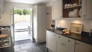 A kitchen with white cabinets, a countertop with a fruit bowl, and a view of a garden through an open door. - No. 2 The Forge, village cottage in idyllic riverside setting (Coniston)