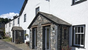 A white building with stone accents, a stone entrance, and a small garden area with potted plants. - No. 2 The Forge, village cottage in idyllic riverside setting (Coniston)