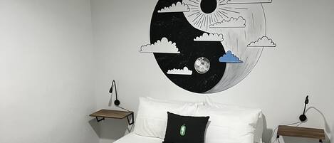 Un lit bien fait, recouvert d’une couverture Nomada noire, avec des oreillers blancs et un coussin noir orné d’un logo vert. Une table de chevet fixée au mur se trouve de chaque côté du lit, et chaque table est surmontée d’une lampe.