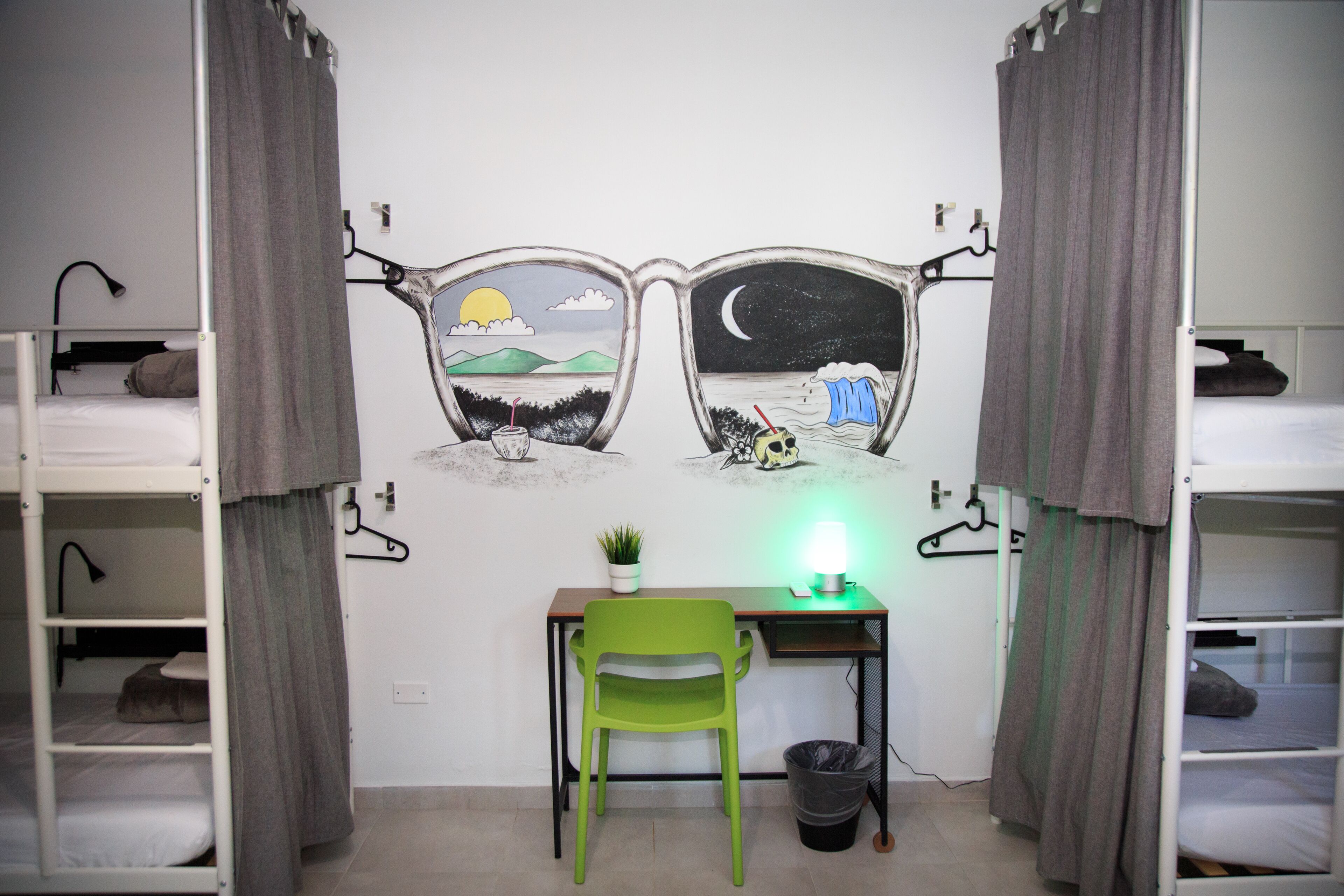 A dormitory room with bunk beds, a desk with a chair, a wall-mounted lamp, and a small plant.