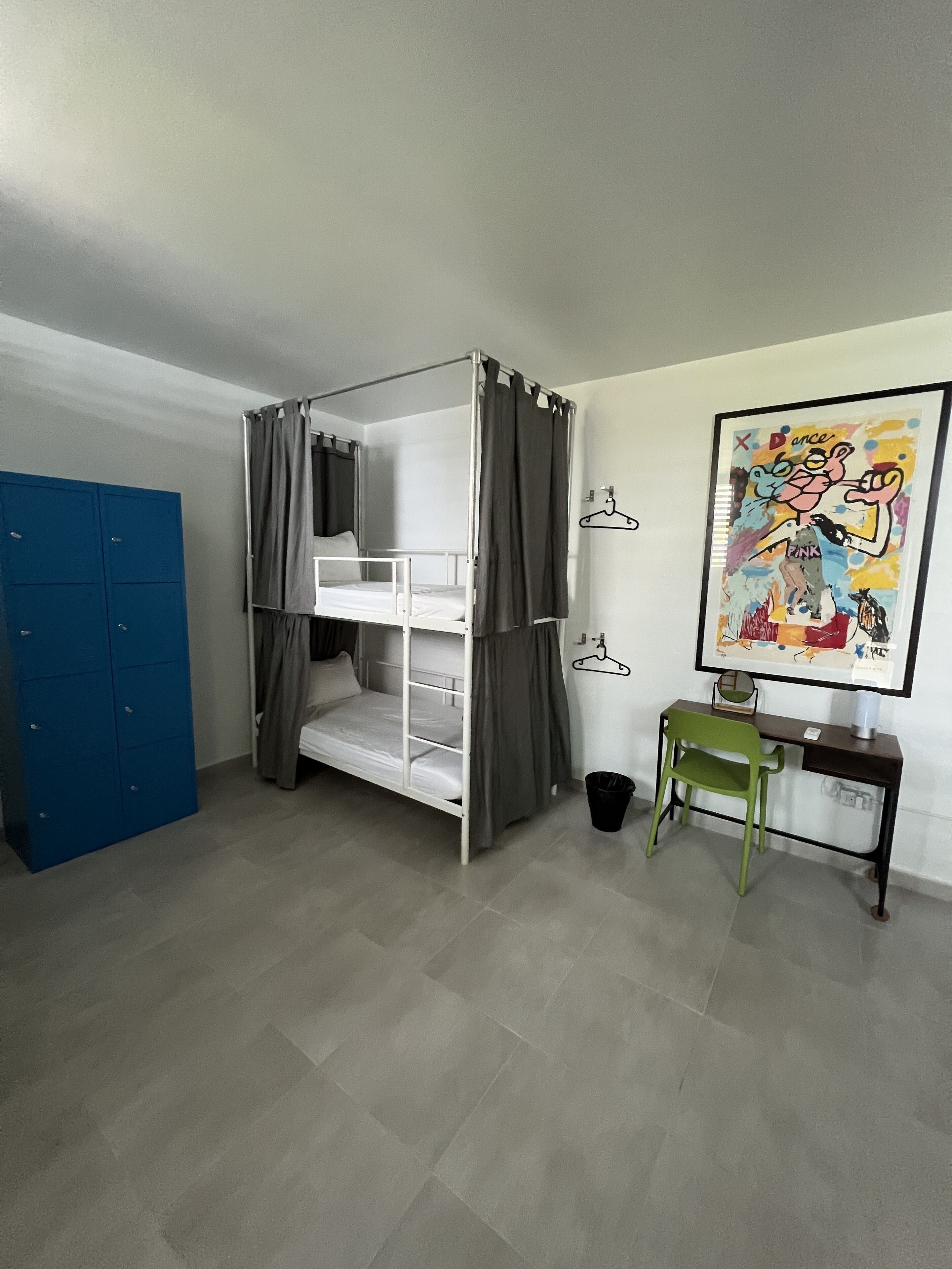 Een kamer met een stapelbed, een bureau, een stoel, een blauwe garderobe en een kleurrijk schilderij aan de muur.