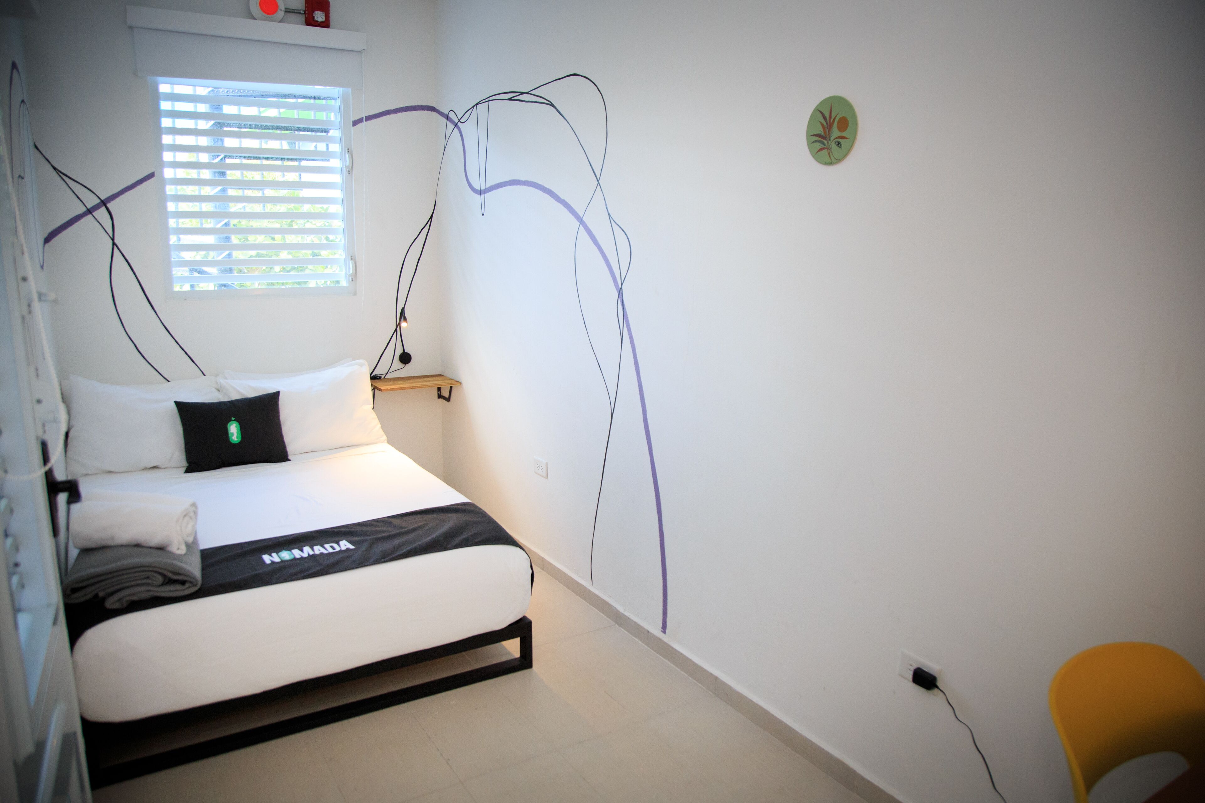 A neatly made bed with a black and white bedspread, a bedside table with a lamp, a window with blinds, and a wall with a decorative plate.