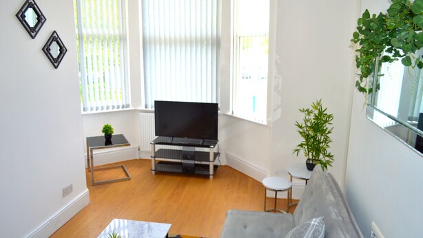 A living room with a flat-screen TV, a grey sofa, a small table with a plant, and a window with blinds. - Beautiful Two-bed Apartment With Driveway Parking (Nottingham)
