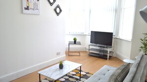 A modern living room with a sofa, a coffee table, a TV stand, and a small table with a plant. - Beautiful Two-bed Apartment With Driveway Parking (Nottingham)