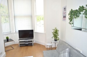 A modern living room with a flat-screen TV, a grey sofa, a small table with a plant, and a window with blinds. - Beautiful Two-bed Apartment With Driveway Parking (Nottingham)