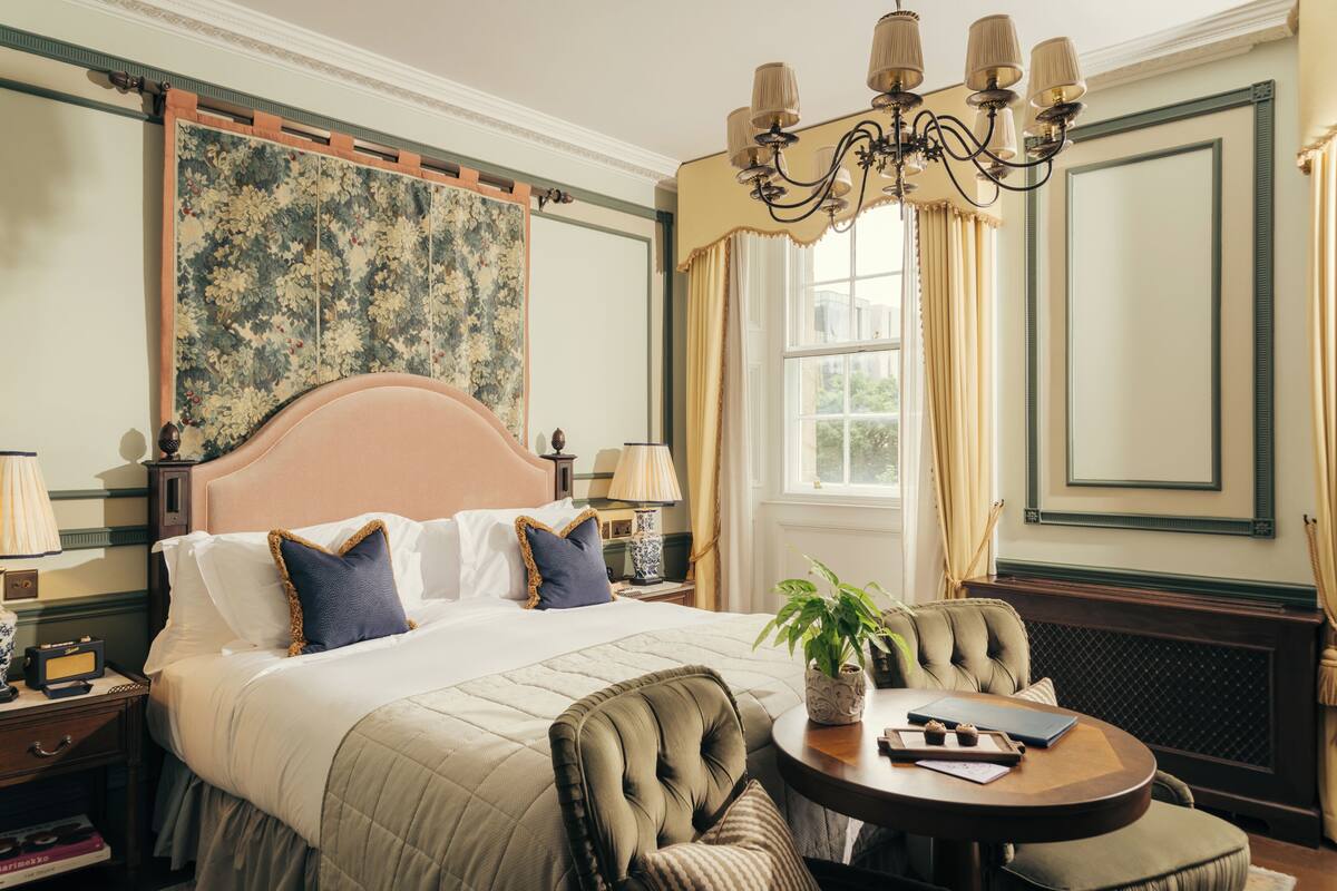 A hotel room with a large bed, a round table, a chair, a nightstand, a lamp, a plant, and a window with curtains.