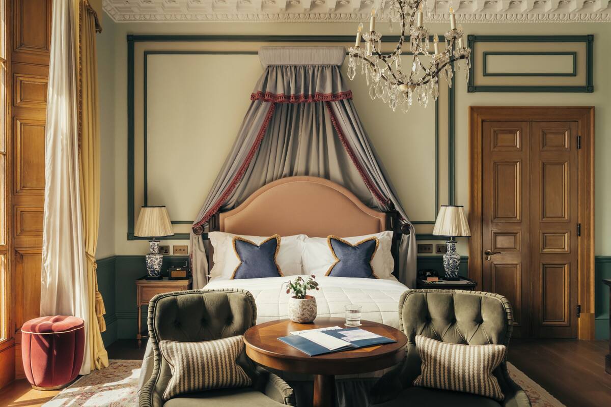 A bedroom with a four-poster bed, two bedside tables with lamps, a round wooden table with chairs, and a chandelier.