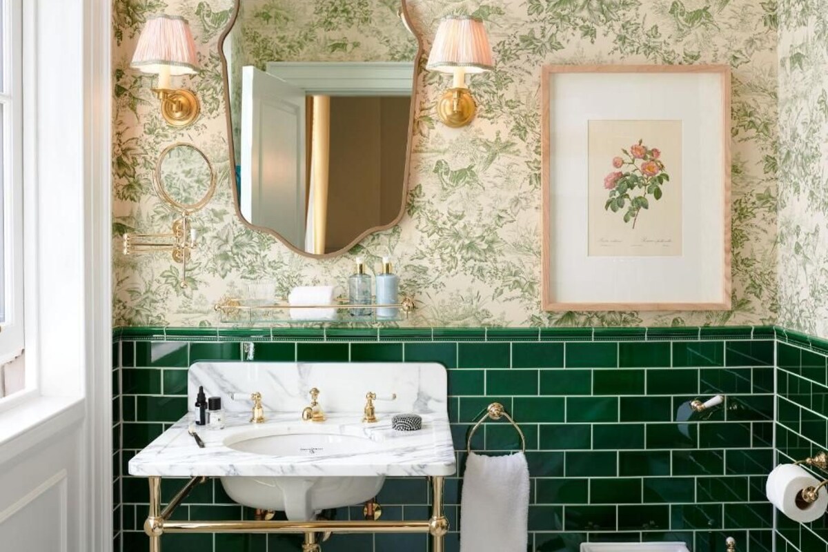 A bathroom with a white marble sink, green tiled walls, a framed floral artwork, and two wall-mounted lamps.