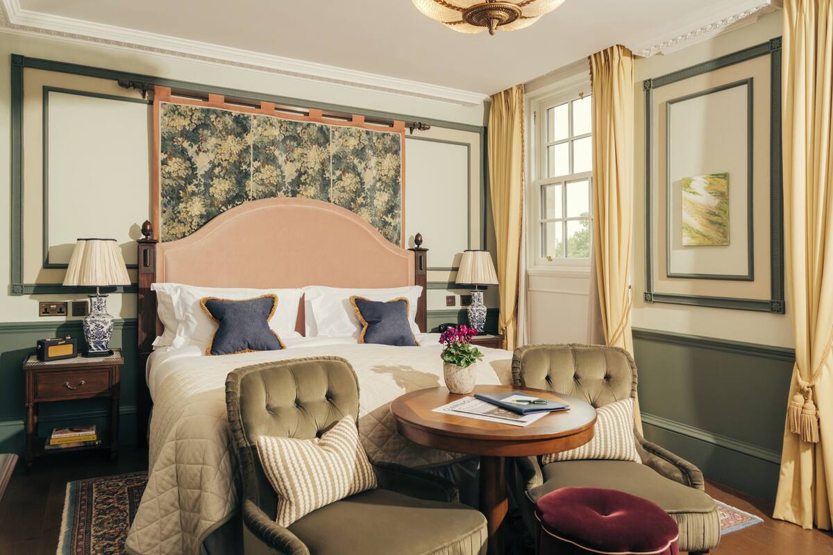 A hotel room with a large bed, two armchairs, a small round table, a nightstand, and a window with curtains.